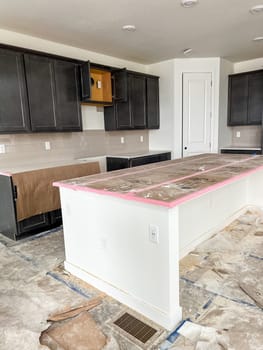 Parker, Colorado, USA-January 11, 2023-A modern kitchen under construction featuring dark cabinets, a large kitchen island wrapped for protection, and unfinished floors. The countertops are partially covered, with construction debris scattered around, showcasing the early stages of a residential kitchen installation.