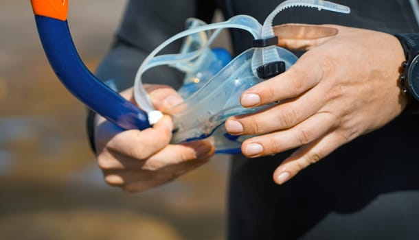 Hands holding a mask and snorkeling tube, a man in a diving suit is preparing to swim and snorkel in the ocean with diving equipment and enjoy the beauty of the underwater world, close-up view.