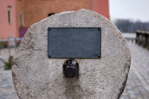 A large stone marker with an empty plaque is set in a riverside area, showcasing its historical significance amid a peaceful urban landscape. Overcast skies add to the tranquil atmosphere.