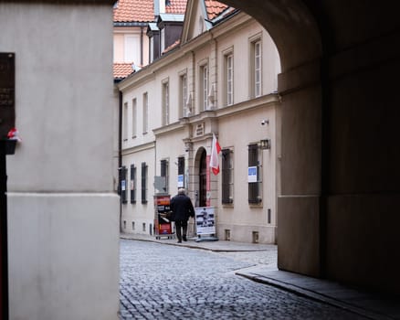 A pedestrian walks down a narrow cobblestone street surrounded by historical buildings. Flags hang from the structures, creating a serene urban atmosphere in a quiet neighborhood.