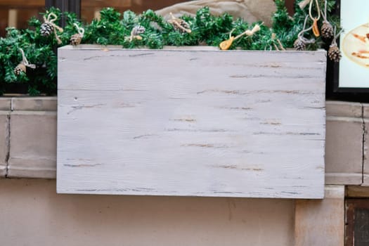 A blank wooden sign is positioned in front of lush greenery and festive decorations, enhancing the inviting atmosphere of a bustling urban restaurant during the holiday season.