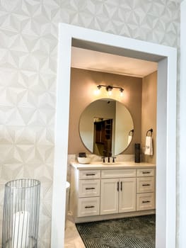Parker, Colorado, USA-January 11, 2023-Stylish bathroom vanity featuring a round mirror, black fixtures, and a bright light fixture above. The vanity has a white countertop with a sleek undermount sink and black faucet. A reflection shows part of a walk-in closet in the background.