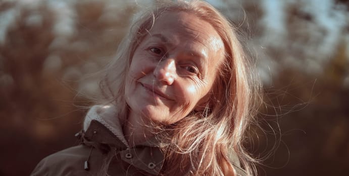 An elderly 60 beautiful woman with a kind smile is walking outdoors on a sunny autumn day, a happy old lady, a grandmother takes care of her appearance, natural beauty with wrinkles portrait closeup.