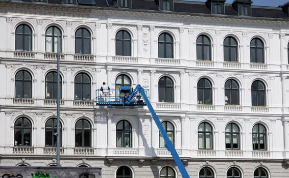 Worker paints the facade of the building at heigt in lifting cradle. Construction workers at height working on crane in lifting bucket. High quality photo