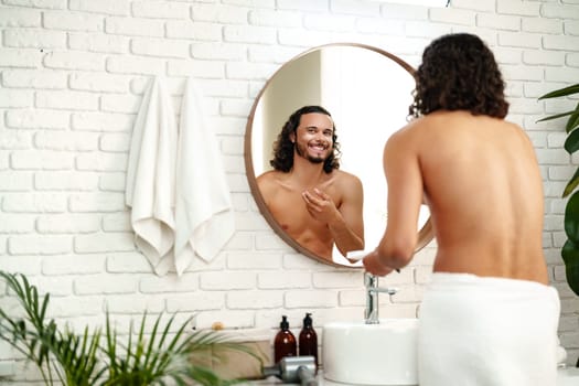 A young man applies skincare cream while looking in a round mirror in a bright bathroom close up