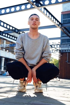 A man wearing a grey sweatshirt, black pants, and white sneakers is squatting on a paved surface. He is looking directly at the camera. There is a large metal framework behind him.