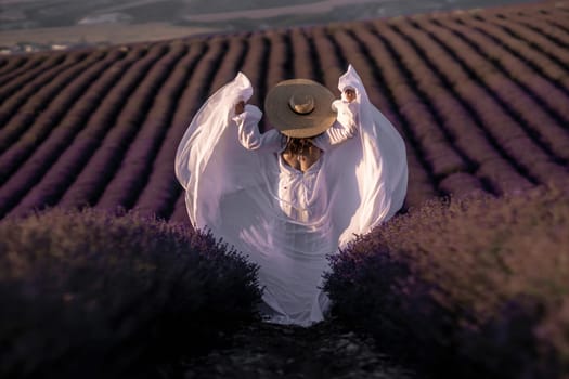 Here's a caption describing the image: Woman in flowing white dress and wide-brimmed hat twirls amidst rows of vibrant purple lavender. Golden hour light bathes the scene.
