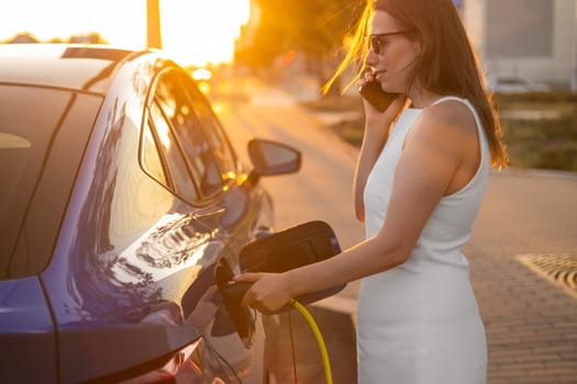 Woman charging an electric vehicle while enjoying a phone call during sunset by the roadside.