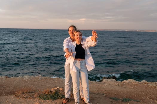 A couple takes a selfie on a rocky shoreline overlooking the ocean at sunset. The man is wearing a white shirt and the woman is wearing a black top and white pants.