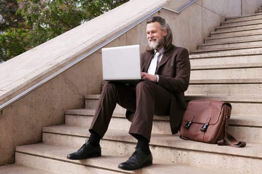 positive mature male entrepreneur in elegant suit siting on urban stairway and working online on laptop in city, concept of business and middle age man lifestyle