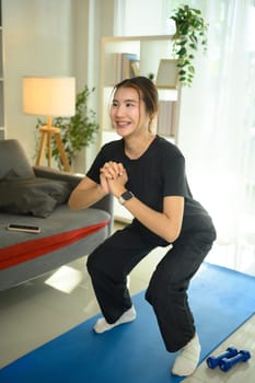 Beautiful young woman doing squat exercise on mat in her living room. Active and healthy lifestyle.