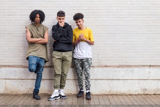 full body of three multiethnic teenager friends with folded hands leaning against white brick wall, concept of friendship in youth and urban lifestyle, copyspace for text