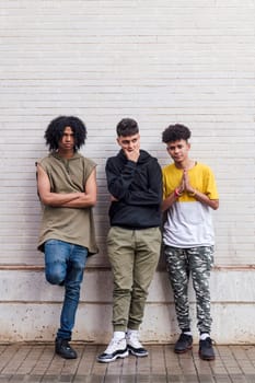full body of multiethnic teenager friends with folded hands leaning against white brick wall, concept of friendship in youth and urban lifestyle, copyspace for text