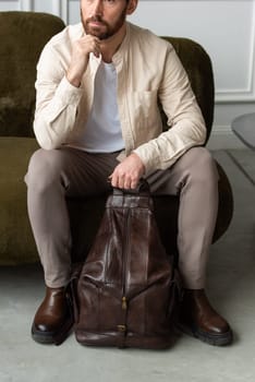 A man is comfortably seated on a sofa with a backpack resting beside him, creating a relaxed and casual atmosphere around him