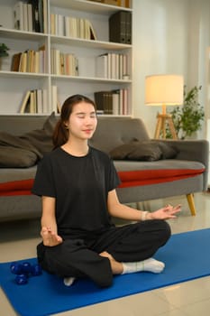 Young woman with eyes closed practicing meditation on a blue exercise mat in living room.