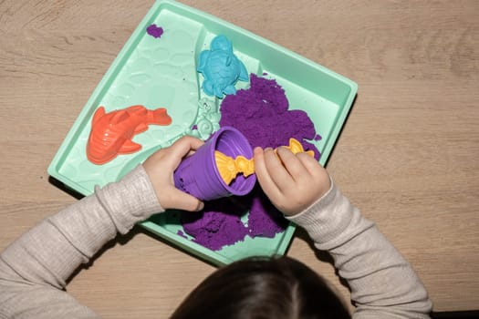 A child is using their fingers to create art with violet and magenta kinetic sand on a table. The leisure activity involves sharing gestures and thumb movements