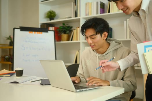 Asian male student in a hoodie working on a laptop, guided by a tutor at home.