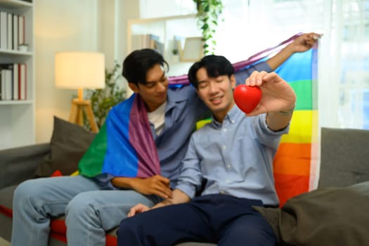 Affectionate gay couple sitting on a couch holding rainbow flag and red heart.