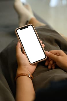 Close up of hands holding a smartphone with white screen while relaxing on sofa.