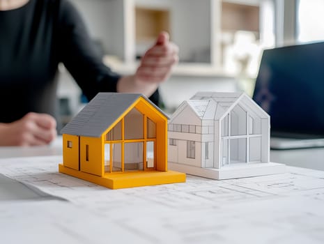 Two detailed house models, one in yellow and one in white, placed on blueprints. A person in the background gestures while discussing architectural designs in a modern office.