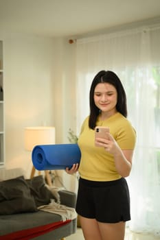 Young sporty woman holding a rolled blue yoga mat and texting message on mobile phone.