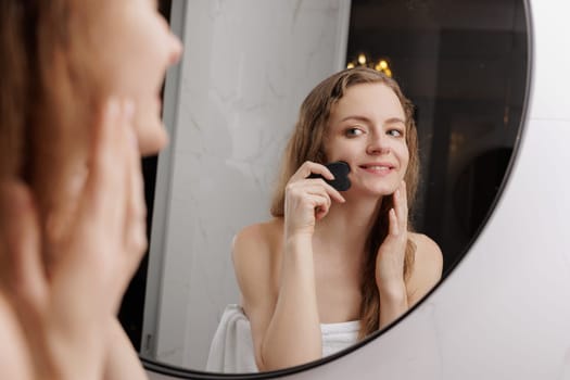 Anti-Aging Skincare. Smiling middle aged woman massaging neck with gua sha tool, attractive mature female making skin lifting treatment with jade guasha stone scraper, standing near mirror at home
