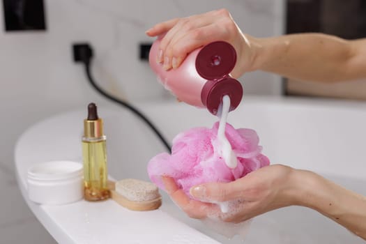 woman's hands applying shower gel on pink washcloth against bathroom background, body and hair care products lying nearby