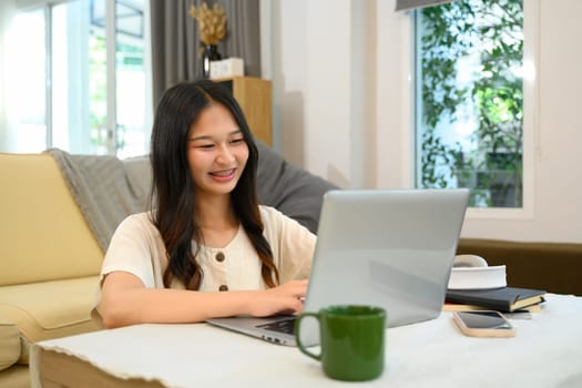 Cheerful young woman freelancer working with laptop on table in living room.