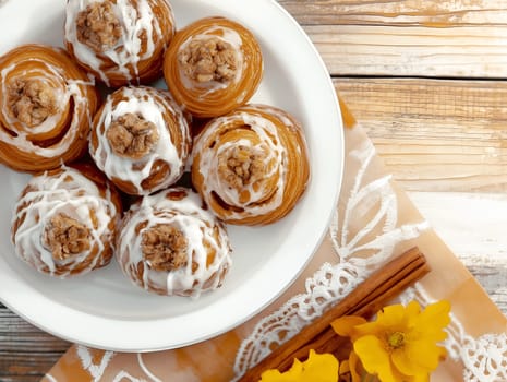 A plate of caramel-glazed cinnamon pastries topped with icing and a walnut center. Styled on a rustic wooden table with yellow flowers and cinnamon sticks for decoration.
