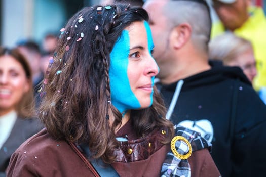 Person at outdoor event with half face creatively painted in bright blue, suggesting celebration or cause, spotted in a vibrant brown jacket.