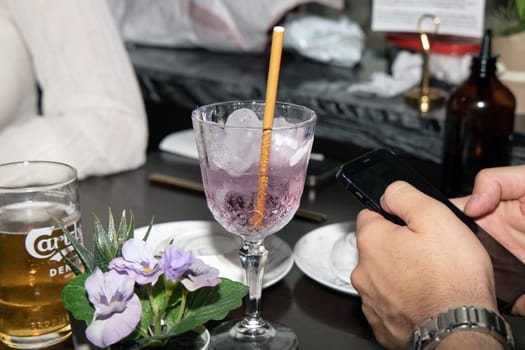 A glass with a lilac cocktail and pieces of ice and beer on a table in a bar, restaurant and men's hands hold a phone. Man and woman in the restaurant. Copenhagen, Denmark - September 4, 2023.