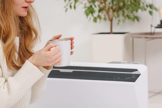 Young woman with warm plaid and tea cup sitting near electric convector heater in bedroom, home heating concept, low temperature