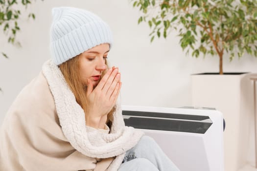 Frozen young woman with plaid warming near radiator at home, home heating concept, low temperature