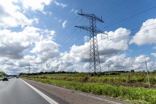 Electricity concept. High voltage power line pylons, electrical tower on a green field with blue sky. Highway with cars on a cloudy summer day.