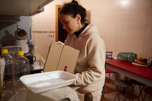 A woman stands in a kitchen holding cardboard boxes, surrounded by various items and utensils.