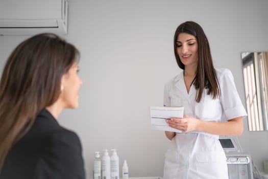 Happy female dermatologist writing prescription while discussing with anonymous client in beauty salon. Beautiful cosmetologist woman is working in clinic.