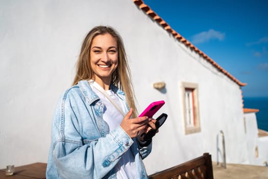 Happy woman holding smartphone while standing at outdoor restaurant during summer. Portrait of smiling attractive female in casualwear by white building.