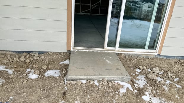 Under-construction backyard patio area featuring a sliding glass door leading to a small concrete step surrounded by dirt and snow. The unfinished landscaping and exposed ground highlight the early stages of residential development.