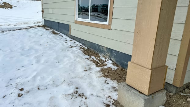 Side view of an under-construction residential home featuring a sliding window framed with trim. The exterior showcases freshly installed siding and an unfinished yard covered with snow. Nearby homes in the background emphasize a suburban development area.