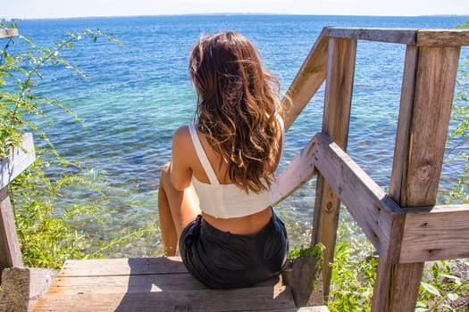 Beautiful young girl with brown hair on the seashore sits and looks at the sea. High quality photo