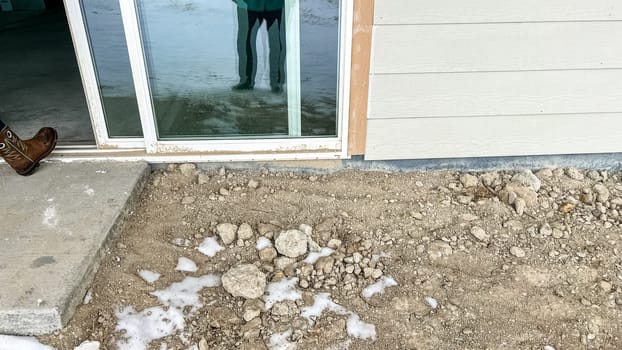 Under-construction backyard patio area featuring a sliding glass door leading to a small concrete step surrounded by dirt and snow. The unfinished landscaping and exposed ground highlight the early stages of residential development.
