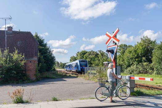 Closed barrier at a railway crossing and a woman cyclist is waiting at a traffic light. A Blue train rides through the countryside. Denmark - Oktober 11, 2023