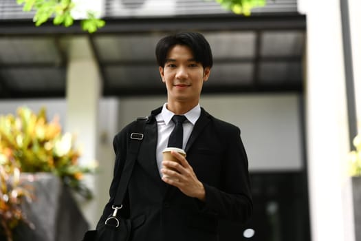 Portrait of young businessman holding takeaway coffee cup walking outside office building. Urban lifestyle concept.