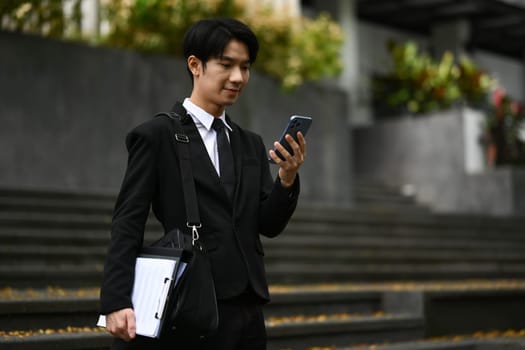 Happy asian businessman texting, checking email on mobile phone during going to work. Technology and city life concept.