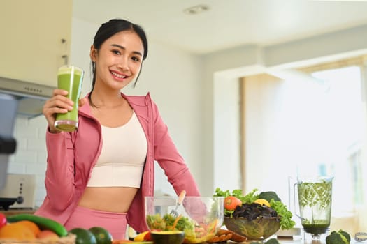 Attractive sports woman holding glass of green smoothie standing in kitchen. Healthy lifestyle concept.