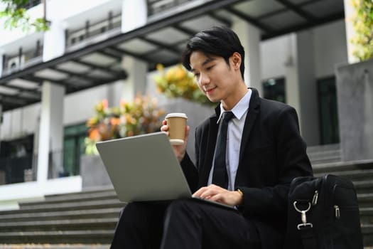 Attractive young businessman sitting on stairs and checking email on laptop before going to the office.