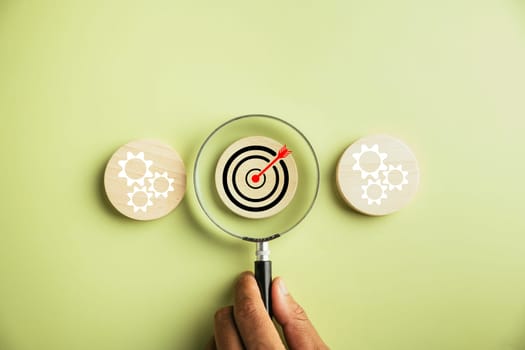 Hand holds a magnifier, focusing on a target icon to analyze objectives and guide management decisions. The image conveys the concept of gear planning, development, and propelling business forward.