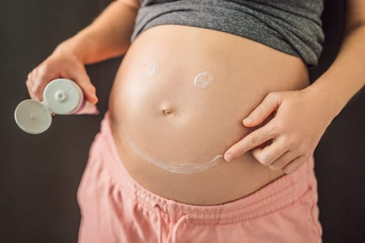 Positive young pregnant woman in comfortable homewear applying belly butter on her big tummy, woman belly.