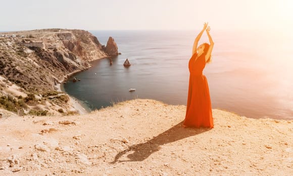 Side view a Young beautiful sensual woman in a red long dress posing on a rock high above the sea during sunrise. Girl on the nature on blue sky background. Fashion photo.