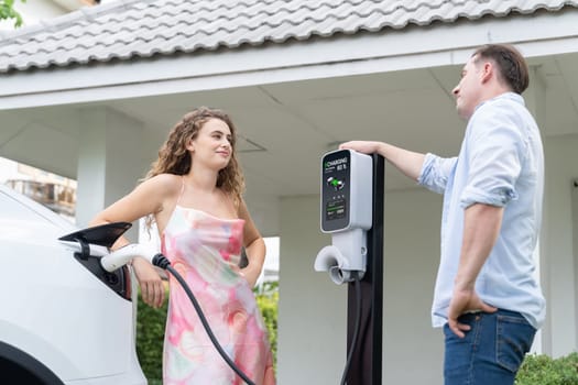 Happy and lovely couple with eco-friendly conscious recharging electric vehicle from EV home charging station. EV car technology utilized for residential home to future sustainability. Synchronos